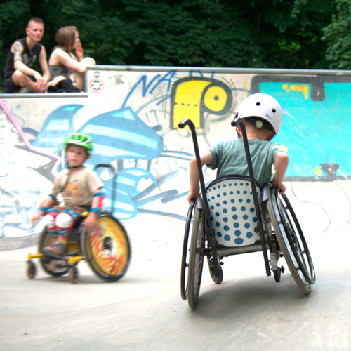 Zwei Jungs mit Helm im Rollstuhl fahren im Skatepark