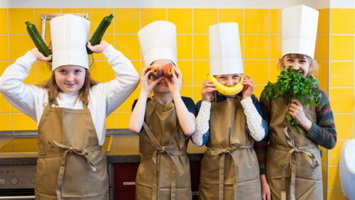 Vier Kinder mit Kochmütze halten sich Gemüse und Obst vor ihr Gesicht. Gesund kochen