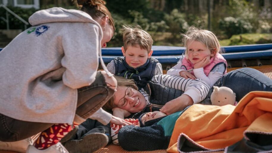 Eine Mutter mit ihren drei Kindern liegen auf einem Trampolin in der Sonne, eine Frau kniet bei Ihnen