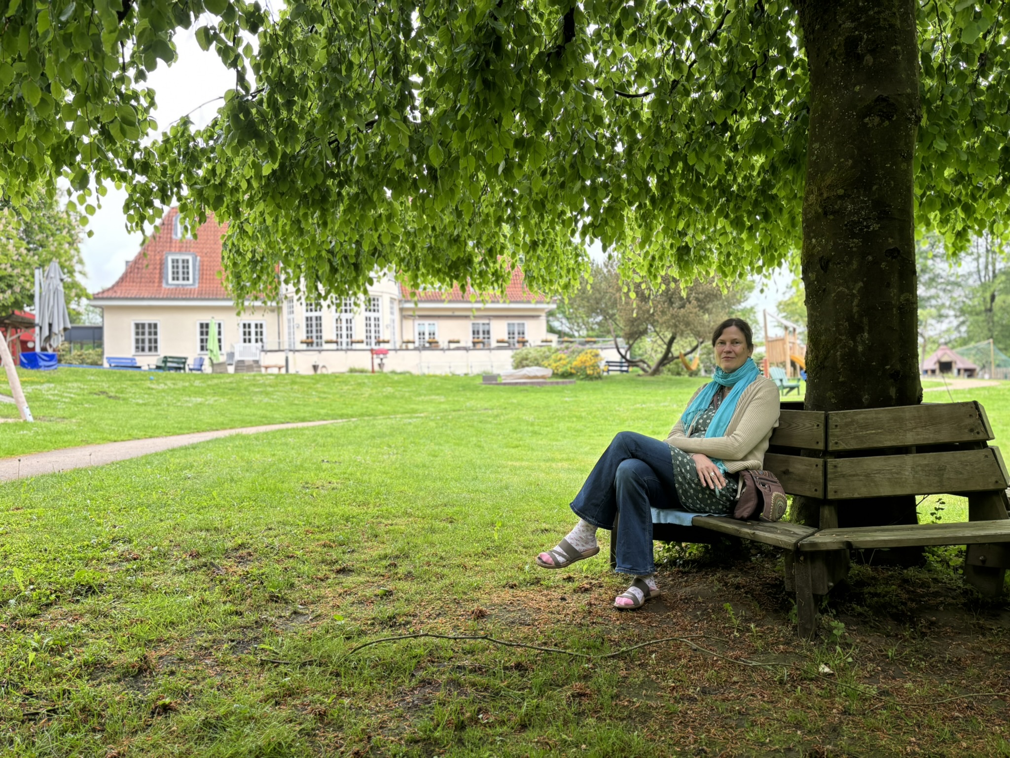 Eine Frau sitzt auf einer Bank unter einem großen Baum im Garten hinter einem Haus.