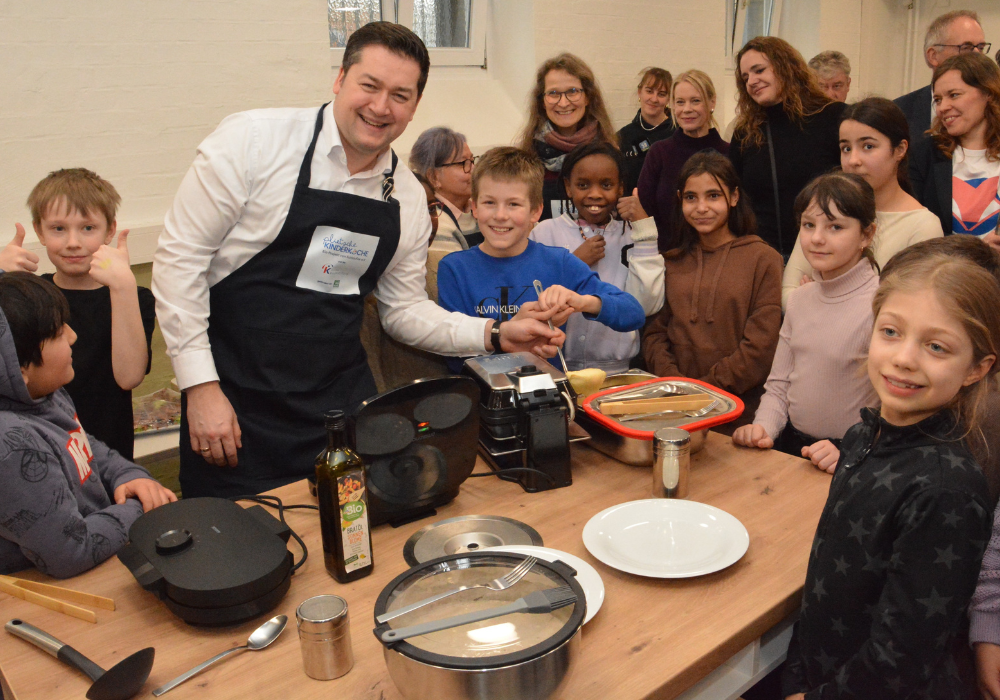 Gruppe großer und kleiner Menschen steht an einem Tisch und löffelt Teig in ein Waffeleisen