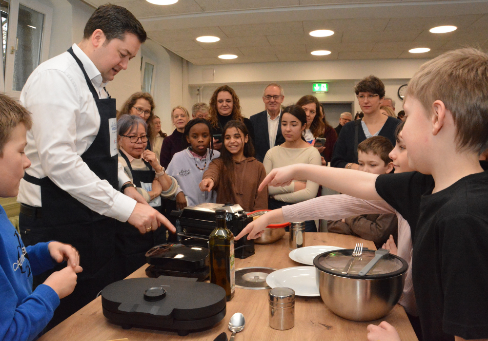 Ein Mann mit Kochschürze steht mit Kindern an einem Tisch und benutzt ein Waffeleisen
