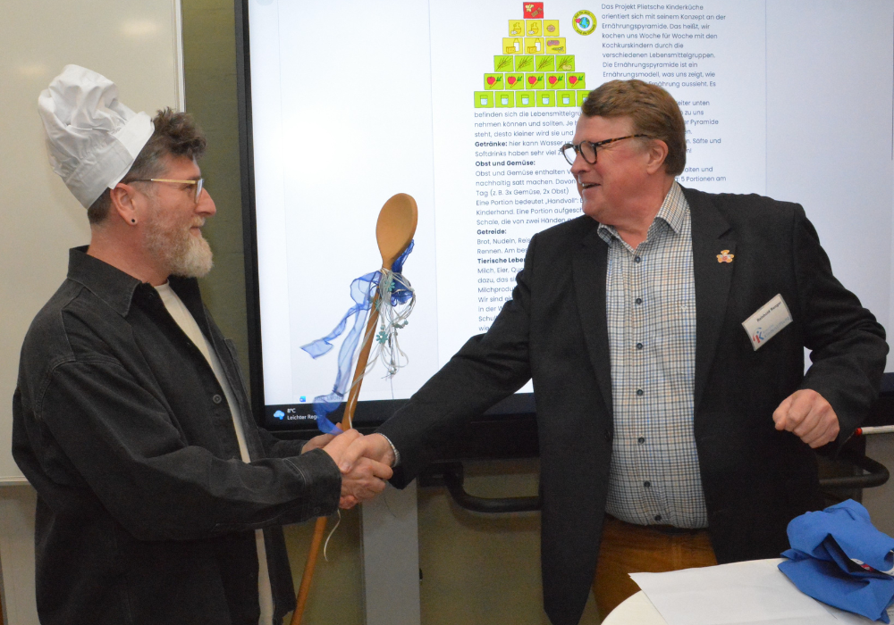 Ein Mann gratuliert einem Anderen, der eine Kochmütze und einen großen Kochlöffel trägt. Sie stehen vor einem Smartboard.