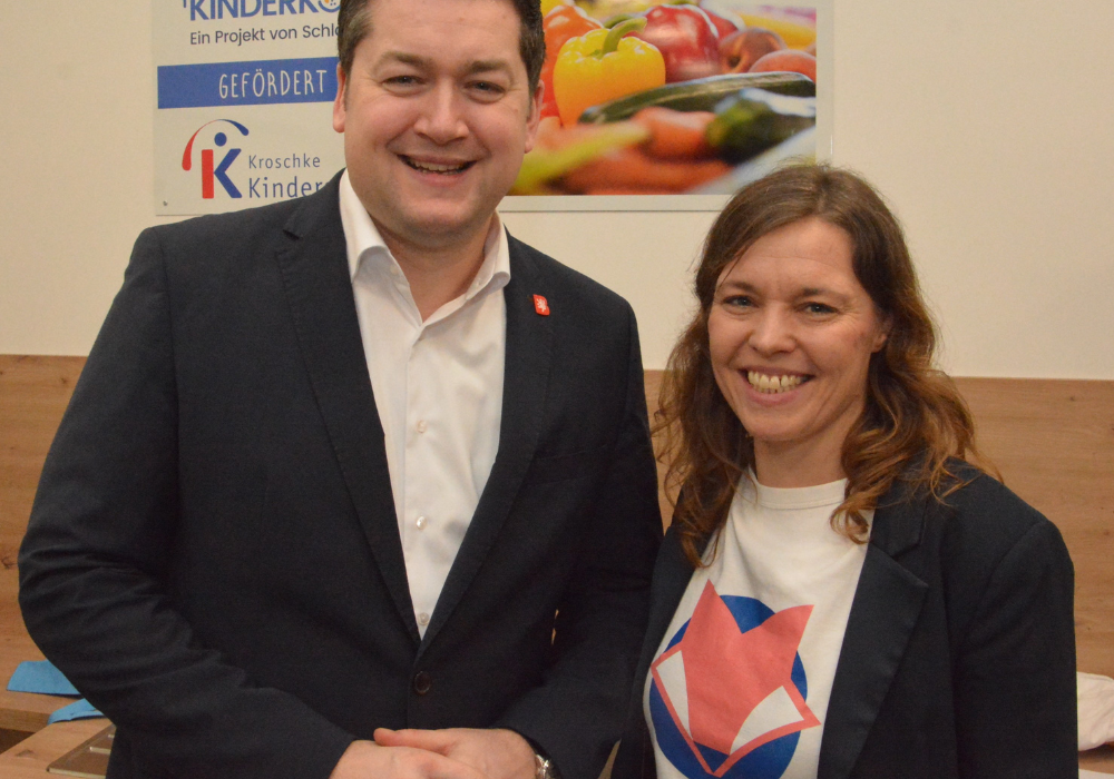 Eine Frau und ein Mann stehen lächelnd vor einem Schild mit der Aufschrift Plietsche Kinderküche