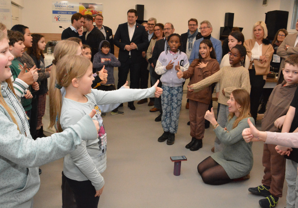 Grundschulkinder stehen im Kreis in einem Raum und singen gemeinsam vor erwachsenen Personen