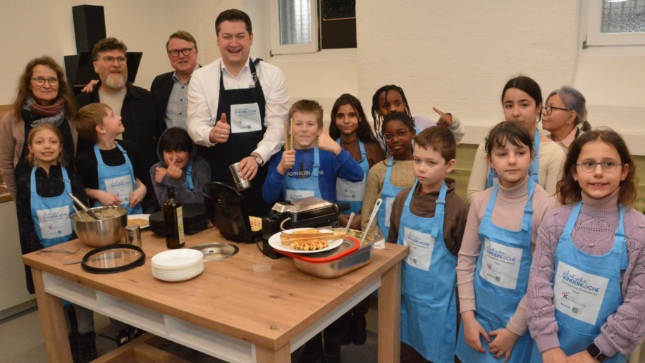 Fünf Erwachsene und elf Kinder stehen um einen Tisch mit Waffeleisen und Waffelteig und bereiten Waffeln zu. Die Kinder und ein Erwachsener tragen Küchenschürzen.