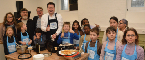 Fünf Erwachsene und elf Kinder stehen um einen Tisch mit Waffeleisen und Waffelteig und bereiten Waffeln zu. Die Kinder und ein Erwachsener tragen Küchenschürzen.