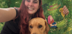 Eine Frau lächelt in die Kamera und hält ein Buch in der Hand. Neben ihr sitzt ein Hund.